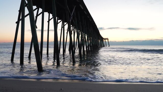 Sunrise viewed beneath Kure Beach pier with sunbeams piercing through wooden pillars.