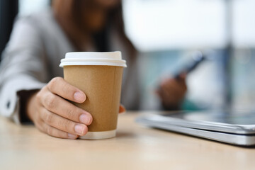Closeup female worker hand holding a paper cup of hot coffee and using mobile phone.