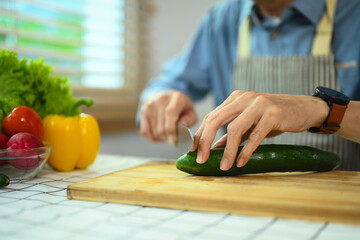 Middle age man cutting cucumber on board preparing wonderful fresh vegan salad in the kitchen.