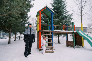 Mom and little girl stand near a snow-covered colorful slide among the trees