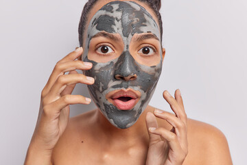 Photo of surprised young Latin woman applies clay mask for skin rejuvenation during her beauty procedures stands shirtless holds breath from amazement isolated over white background. Wellness concept