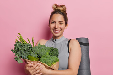 Balanced and nourishing lifestyle concept. Positive fit woman with hair gathered in bun holds paper bag full of fresh green vegetables rubber fitness mat smiles happily isolated over pink background - obrazy, fototapety, plakaty
