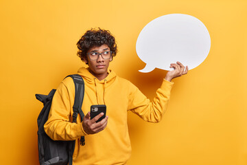 Horizontal shot of thoughtful curly haired Hindu man holds blank speech bubble and modern smartphone for online communication wears spectacles and hoodie carries rucksack isolated on yellow background