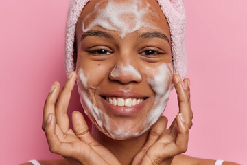 Beauty and cosmetology concept. Studio shot of young pretty smiling happy European lady applying soap on face as part of daily routine procedures holding hands near cheeks posing on pink background - obrazy, fototapety, plakaty
