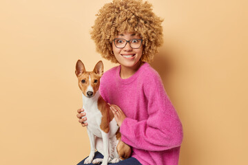 Curly haired woman shares cozy moment with her jack russell terrier dog poses on chair enjoys heartwarming bond bites lips from curiousity wears spectacles and pink jumper isolated over brown wall