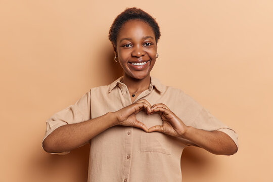 Studio Portrait Of Young Delighted Smiling Broadly African American Woman Stading In Centre Isolated On Beige Background Showing Love To People Making Heart With Hands Expressing Positive Emotions