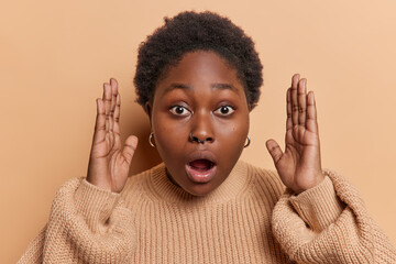 Big size concept. Scared dark skinned plump African woman with curly hair holds breath keeps mouth opened shows something very big dressed in casual knitted jumper isolated over brown background