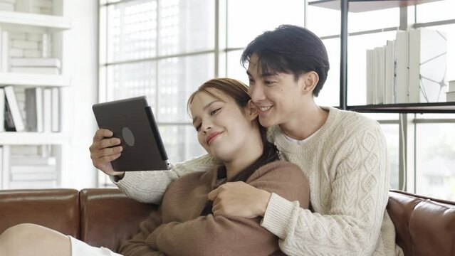 Happy asian young couple looking at digital computer tablet screen, relaxing on comfortable couch. Loving couple shopping online in internet store, watching media content online