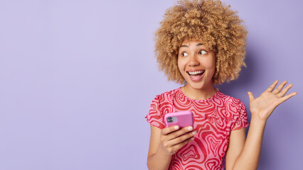 People positive emotions concept. Indoor photo of young pretty cheerful smiling African american lady standing on right isolated on purple background holding smartphone with blank space for promotion