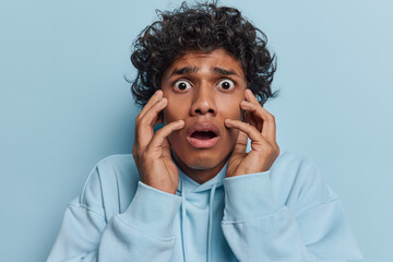 Photo of frightened Hindu man with curly hair keeps hands on cheeks reacts to something shocking trembles from fear wears casual hoodie isolated over blue background. People and reactions concept - obrazy, fototapety, plakaty
