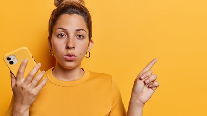 Horizontal shot of surprised woman with hair bun points index finger at empty space for your...