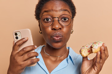 Horizontal shot of dark skinned curly woman keeps lips rounded holds mobile phone and delicious...