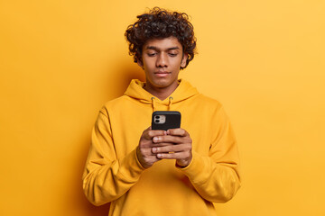 Serious curly haired Hindu man holds smartphone in hands reads news online browsing website...