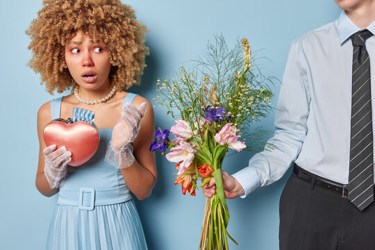 Shocked curly haired woman with bouquet of flowers in hand her boyfriend expresses his love despite being aware of her allergies wears elegant clothing looks worried isolated over blue background
