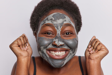 Close up shot of short haired woman with dark skin applies facial clay mask for skin rejuvenation clenches fists and smiles broadly feels very happy isolated over white background. Beauty procedures