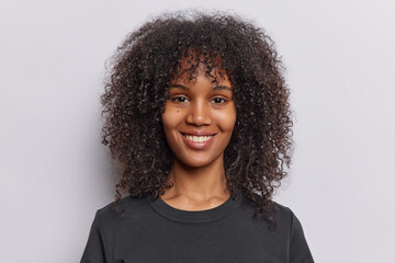 Portrait of cheerful curly haired teenage girl with healthy skin perfect teeth dressed in casual black t shirt looks directly at camera isolated over white background. People and positive emotions