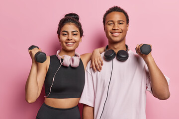 Sporty motivated woman and man lift dumbbells for training muscles do exercises in gym dressed in sportswear use headphones for listening music isolated over pink background. Healthy lifestyle concept