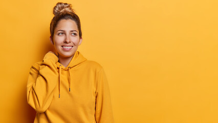 People positive emotions concept. Indoor shot of young pretty glad smiling European lady standing on left isolated on yellow background wearing hoodie looking aside with blank space for advertisement