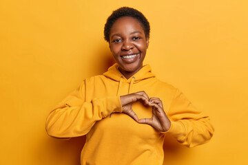 Lovely dark skinned woman with short curly hair smiles broadly dressed in casual sweatshirt expresses love makes heart gesture has romantic mood isolated over yellow background. Be my valentine