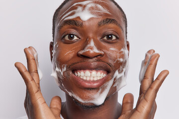 Close up shot of cheerful dark skinned man applies cleansing gel on face has fun whille washing...