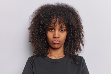 Portrait of upset curly haired woman purses lips has disappointed face being discontent by something wears black t shirt isolated over white background. People and negative emotions concept. - obrazy, fototapety, plakaty