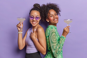 Positive young pretty women stand back to each other have cheerful expressions hold cocktails enjoys party time and celebration feel carefree wear stylish clothing isolated over purple background - Powered by Adobe