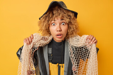 Shocked curly haired fisherwoman stands shocked holding fishing net has passion for sea just returning from successful catch stunned to catch big fish isolated over yellow background. Hobby concept