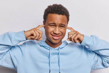 Displeased African man with sulking expression plugs ears has unhappy expression wears casual blue...