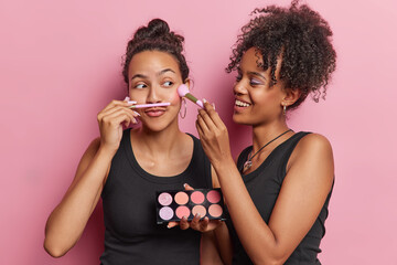 Waist up shot of cheerful two young women apply colorful eyeshaow with cosmetic brushes going to do makeup dressed in black t shirts stand closely to each other isolated over pink background