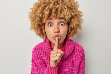 Stunned curly haired woman keeps index finger over lips telling secret shares confidential...