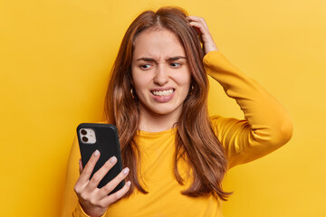 Displeased clueless young woman keeps hand on head clenches teeth and focused at smartphone dressed...