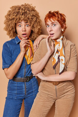 Vertical shot of two surprised women dressed in fashionable clothing cannot believe in something shocking have widely opened eyes and mouthes stands next to each other isolated over brown background