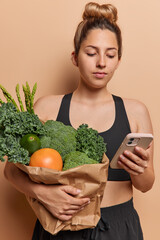 Vertical image of sporty European woman dressed in black tracksuit poses with paper bag of green vegetables concentrated in smartphone finds diet for slimming isolated over brown background.