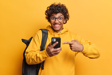 Curly haired Hindu man with glad expression points index finger at smartphone suggests to download...
