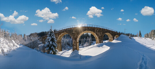 Stone viaduct (arch bridge) on railway through mountain snowy fir forest. Snow drifts  on wayside and hoarfrost on trees and sunshine in sky.