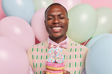 Photo of dark skinned man celebrates anniversary poses with doughnuts and burning candles dressed...