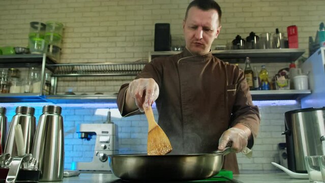 Professional chef standing in well-equipped kitchen stirs searing dish using spatula mixing flavors of ingredients. Male cooking concept