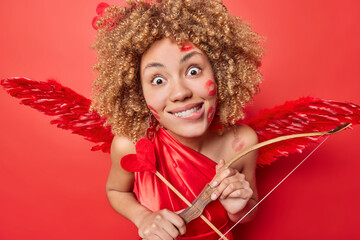 Horizontal shot of curly haired European woman looks surprisingly at camera bites lips holds arrow...