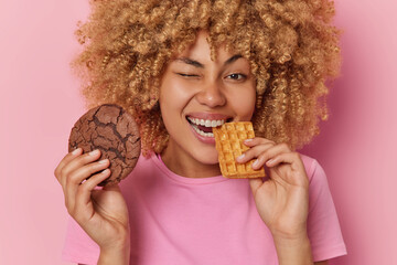 Positive curly haired young woman eats delicous cookie and waffle winks eye and smiles broadly...