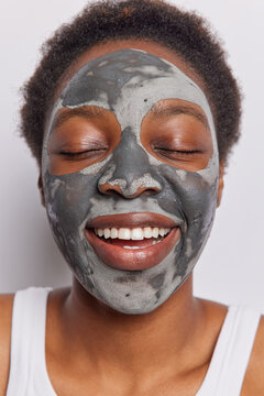 Close up shot of cheerful African woman keeps eyes closed applies facial clay mask for reducing wrinkles and pores dressed in casual t shirt isolated over white background. Face treatments concept