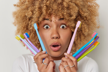 Headshot of curly haired speechless woman with blonde curly hair holds colorful toothbrushes chooses best one to clean teeth stares with omg expression at camera poses against white background.