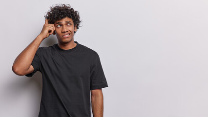 Waist up shot of confused curly haired young Hindu man scratches head and looks pensively aside thinks about something wears casual black t shirt isolated over white background. Hmm let me think - 701435772