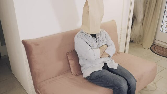 A man wearing a breadbag face, sitting on the sofa at home, no expression on his paper mask; doing nothing, feeling bored.
