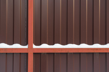 Fragment of a metal fence made of brown profiled sheet. There are frame elements. There is a wavy layer of snow on a horizontal beam. Background. Form.