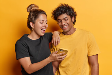 Two positive friends watch funny video in internet look at screen of smartphone happily have broad smiles on faces stand closely to each other dressed in casual t shirts isolated on yellow background - 701437305
