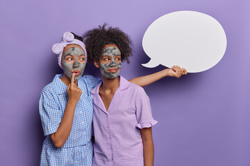 Thoughtful young women with purifying mask on face concentrated aside holds blank speech bubble for your advertising content dressed in nightwear isolated over purple background. Studio shot