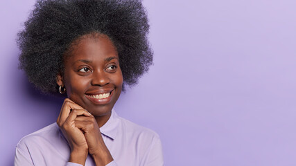 Studio shot of pretty dark skinned lady with bushy curly hair keeps hands near face concentratedd aside has gentle smile dreamy expression dressed formally isolated over purple background copy space