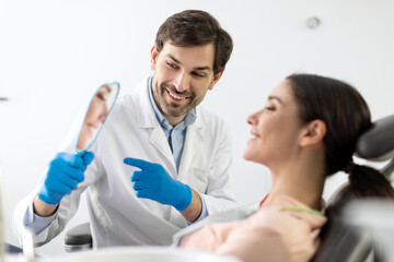 Friendly professional male dentist giving mirror to his happy female patient, showing result of dental treatment or whitening