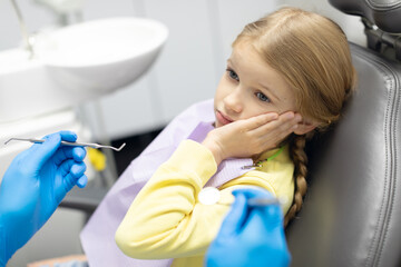 Sad child girl sitting in dentist's chair and holding hand to his cheek from toothache and looking...