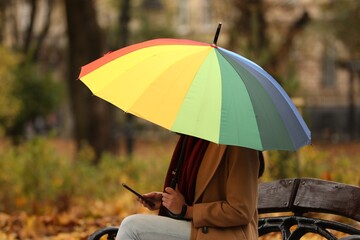 Woman with rainbow umbrella and smartphone sitting on bench in autumn park